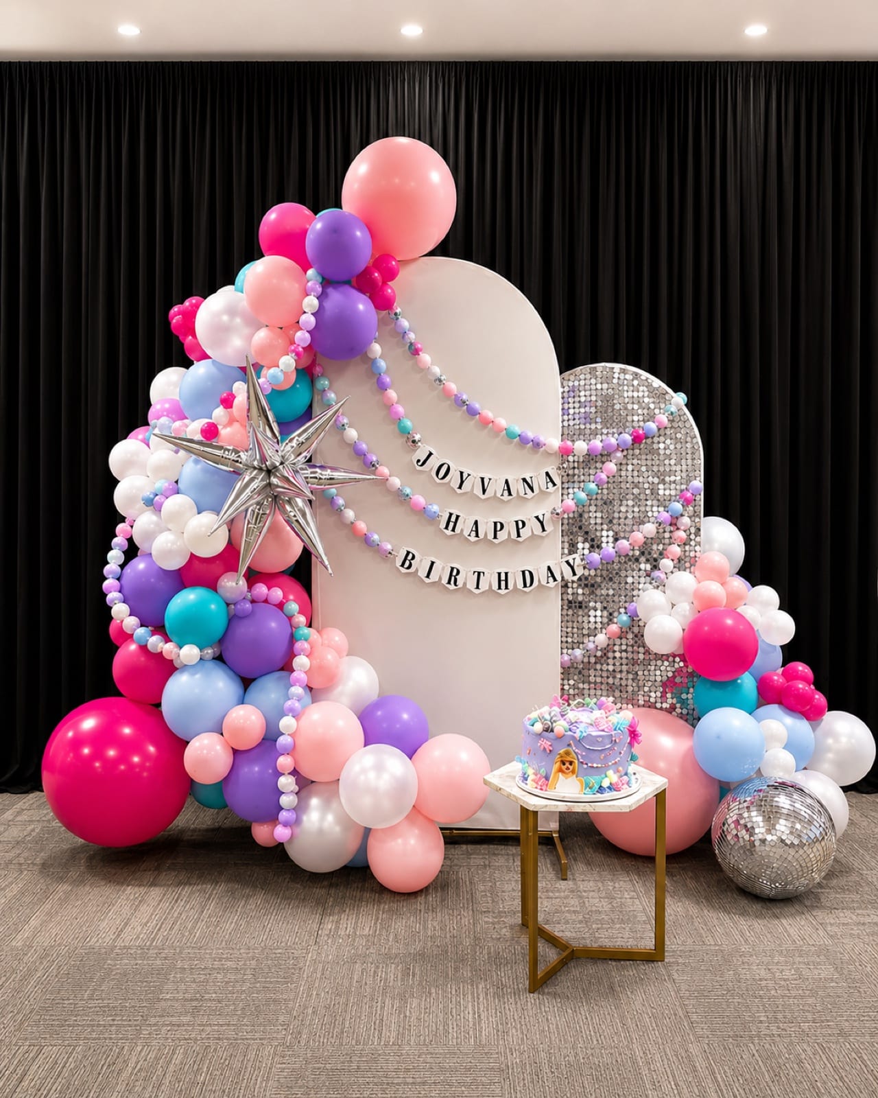 Pink, violet and sky-blue balloons banked around a white arch and a silver sequin wall