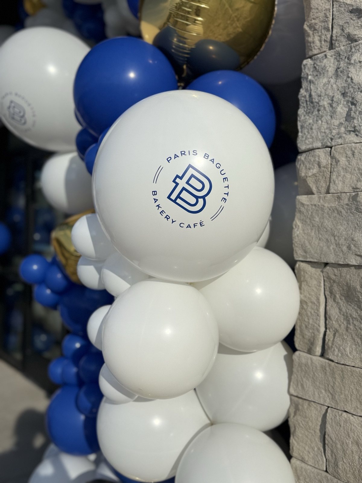 A white balloon printed with a bakery café’s navy logo, clustered with royal blue, white and gold balloons at a grand opening