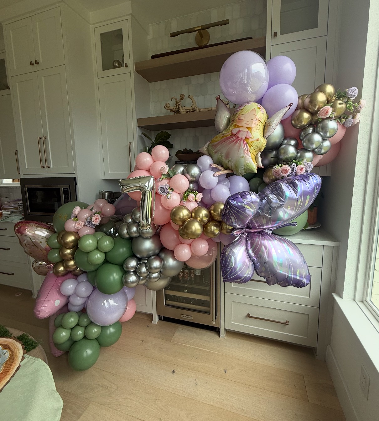 A sage, blush and lilac garland threaded with chrome and gold around a kitchen bar, hung with butterfly foils, a fairy and a silver number one