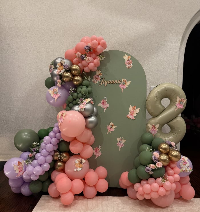 A sage, coral and lilac garland threaded with chrome, climbing an arched backdrop scattered with paper fairies beside a number eight