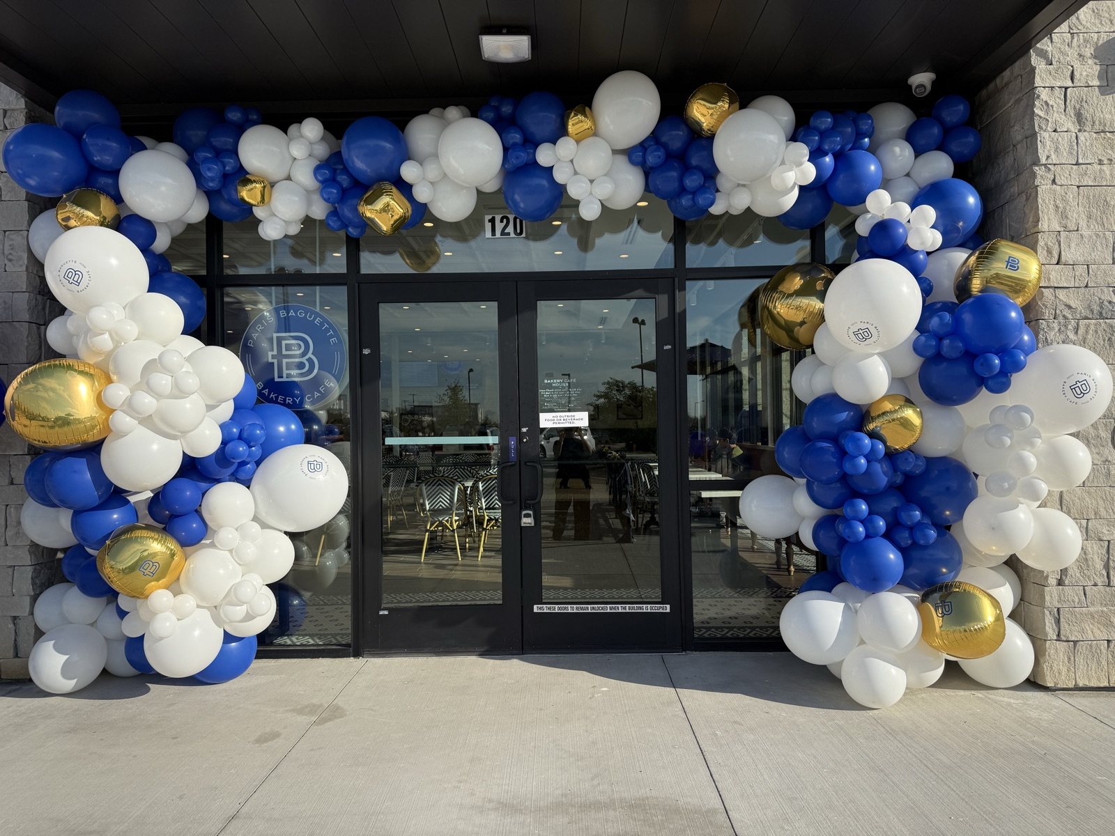 An organic arch of royal blue, white and gold balloons framing a bakery’s glass entrance, the white balloons printed with the café’s logo
