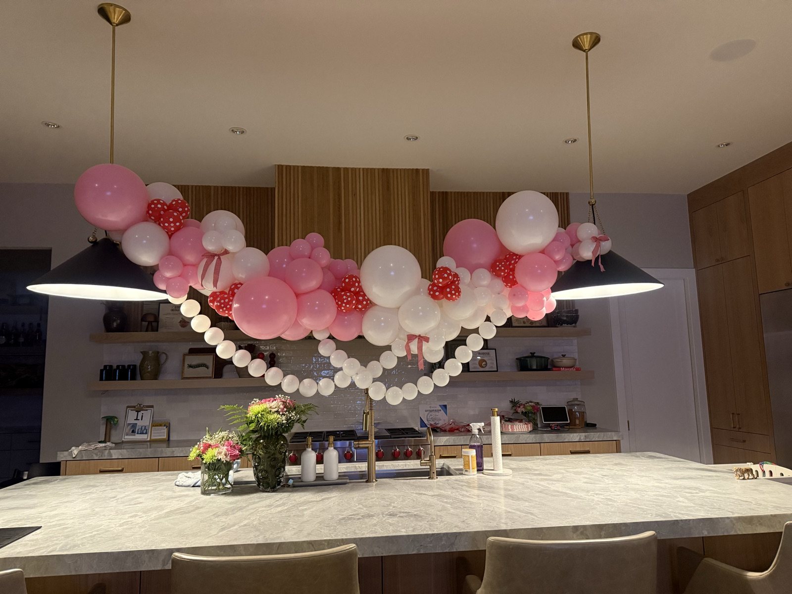 A swag of blush, pink and white balloons with red polka-dot hearts strung between two black pendant lamps over a marble kitchen island