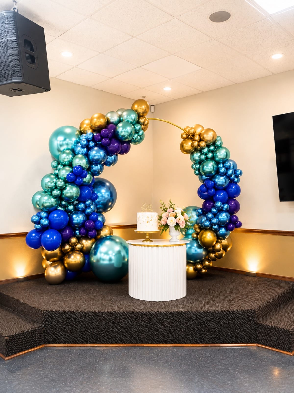 A ring arch of chrome balloons in sapphire, teal, violet and gold curving around a cake table