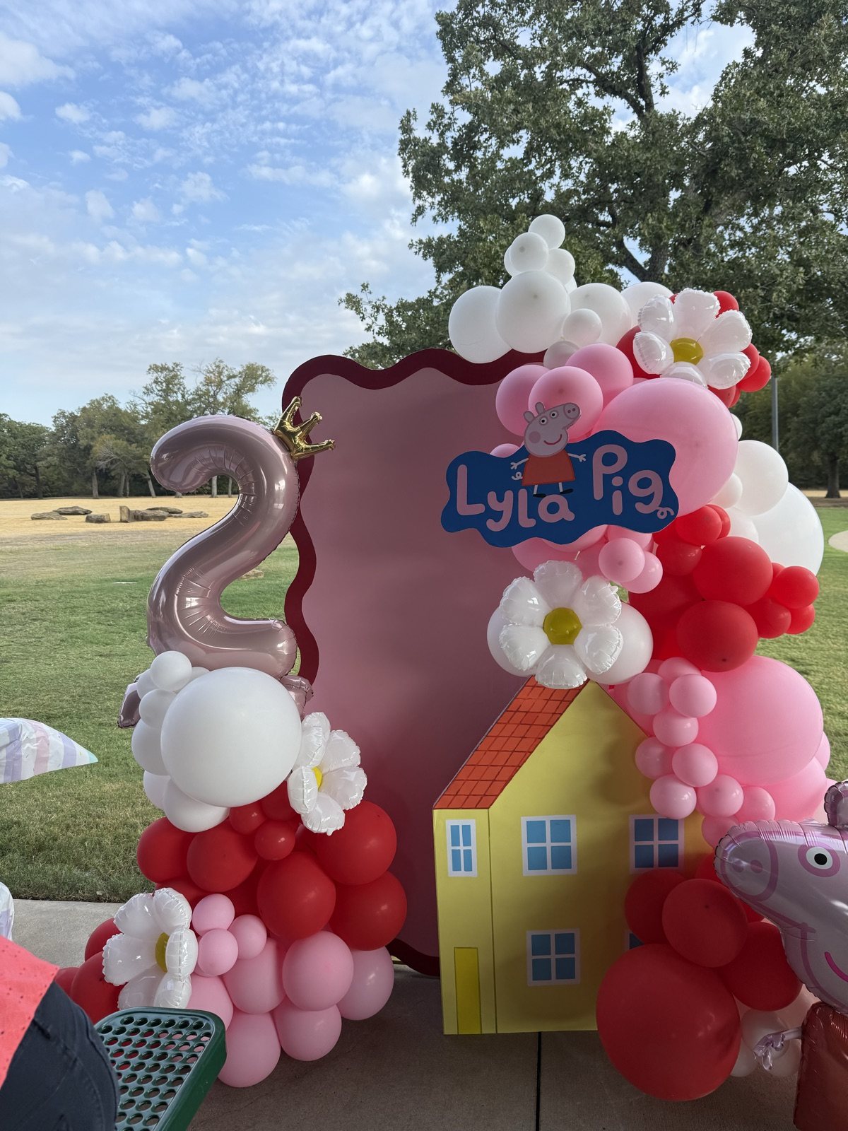 Red, pink and white balloons with daisy foils around a wavy pink backdrop and a yellow cut-out house, beside a rose-gold number two with a crown
